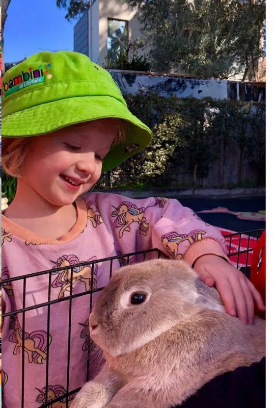 A child in a green Bambini hat smiling beside a grey rabbit during an animal visit to the centre