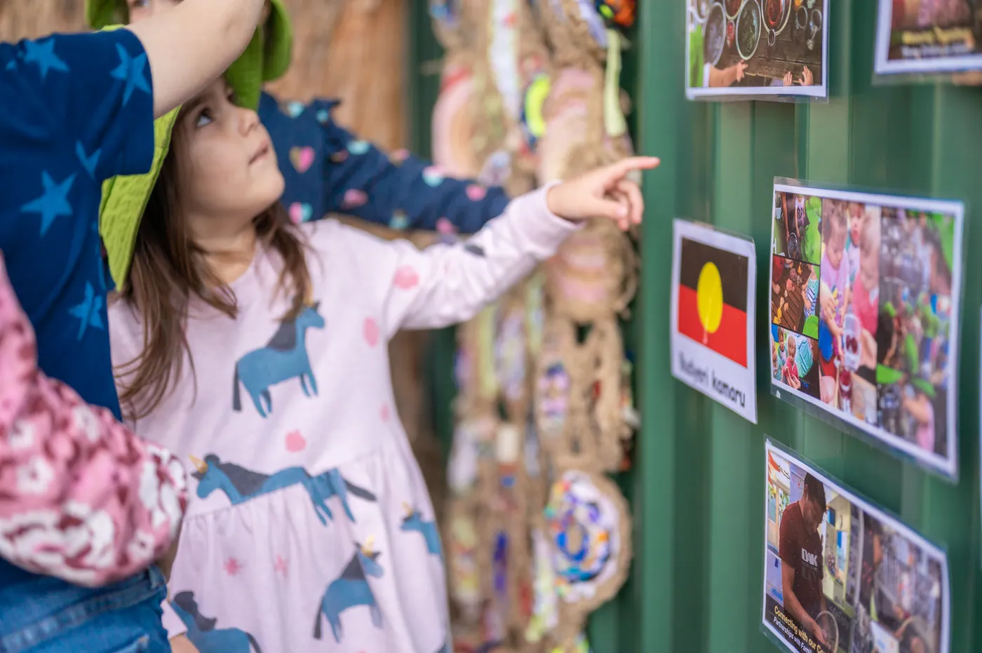 A child in a Bambini hat pointing up at photographs pinned beside a tall woven wall hanging