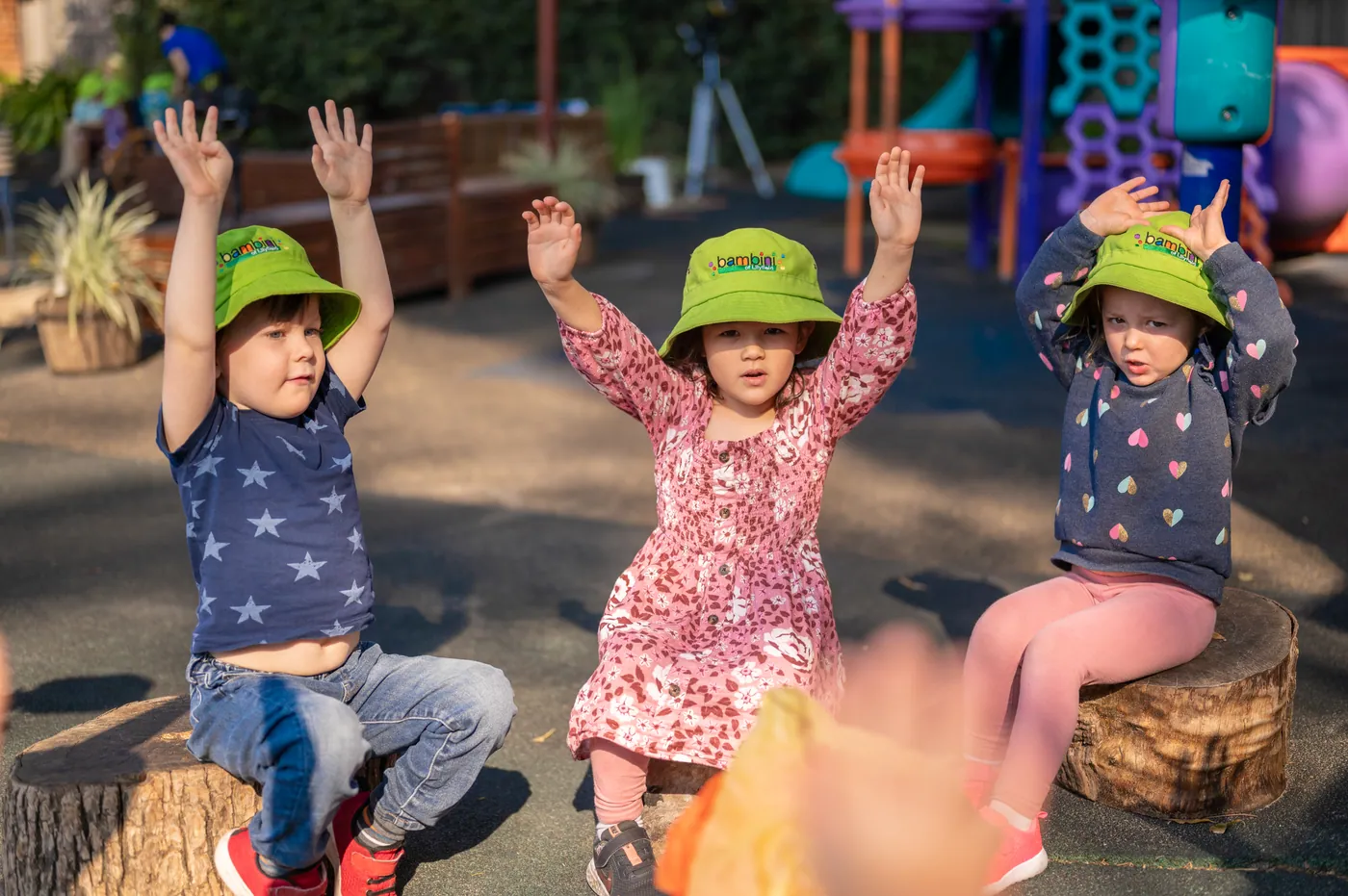 Three children sitting on tree stumps outdoors with their arms in the air
