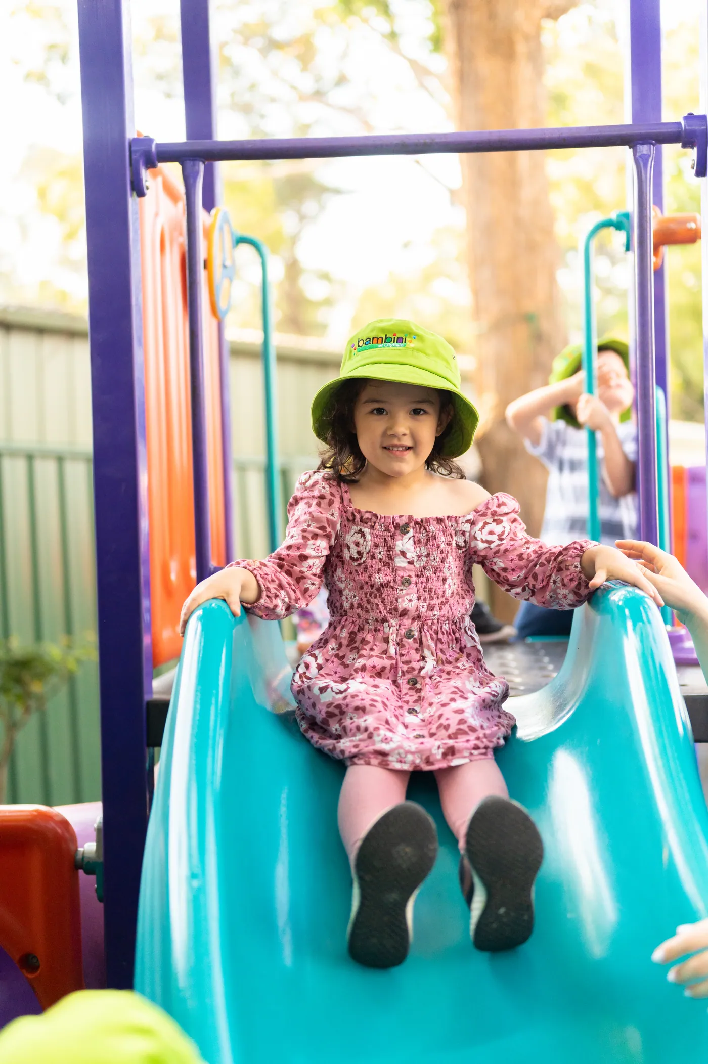 A child in a green Bambini hat sitting at the top of the turquoise slide, ready to push off