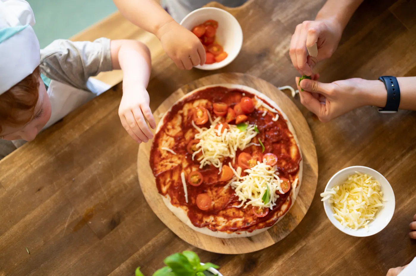 A child in a paper chef's hat adding cherry tomatoes to a pizza base while an educator tears basil over it