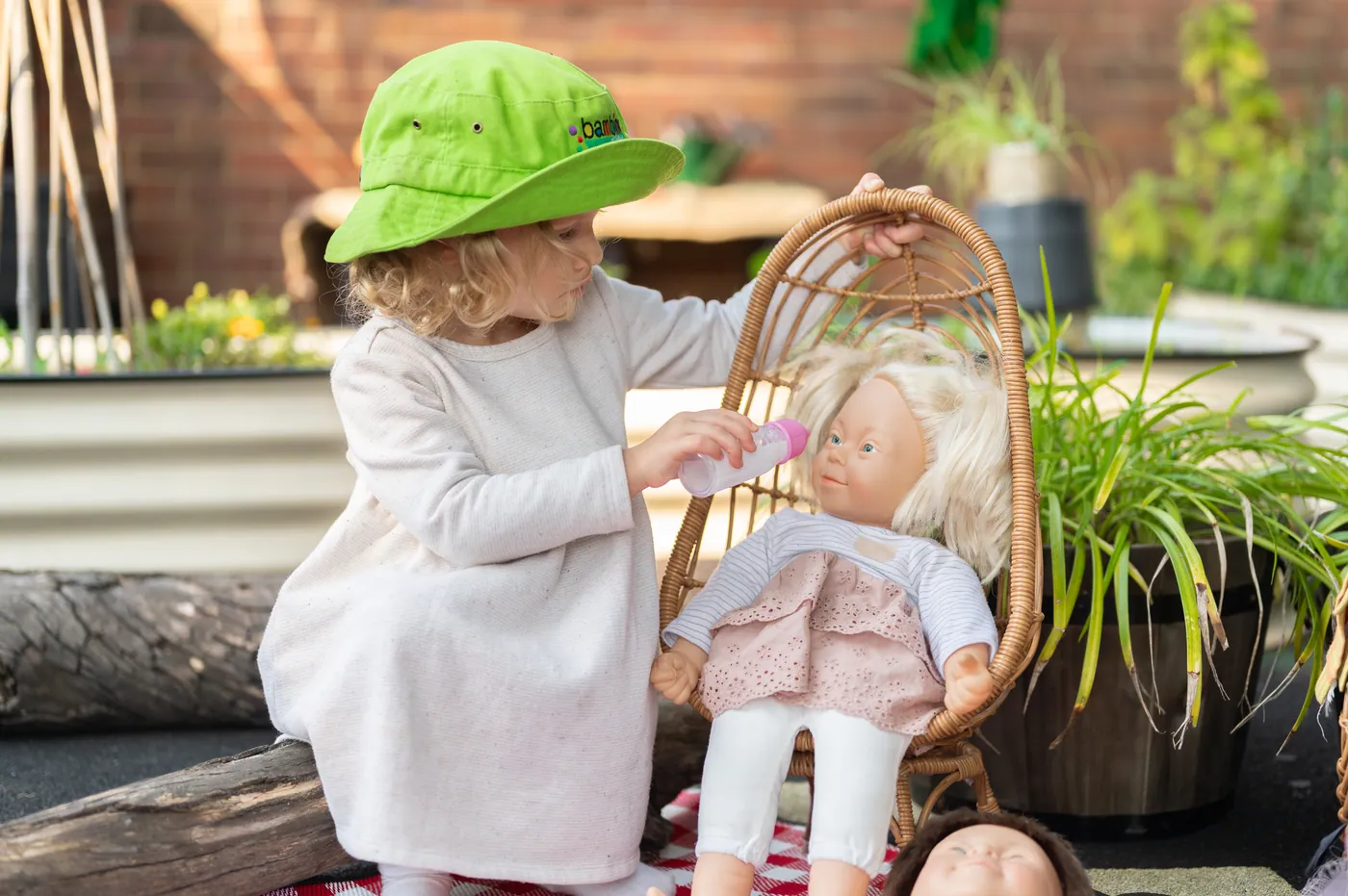 A child in a green Bambini hat holding a toy bottle to a doll propped up in a small cane chair