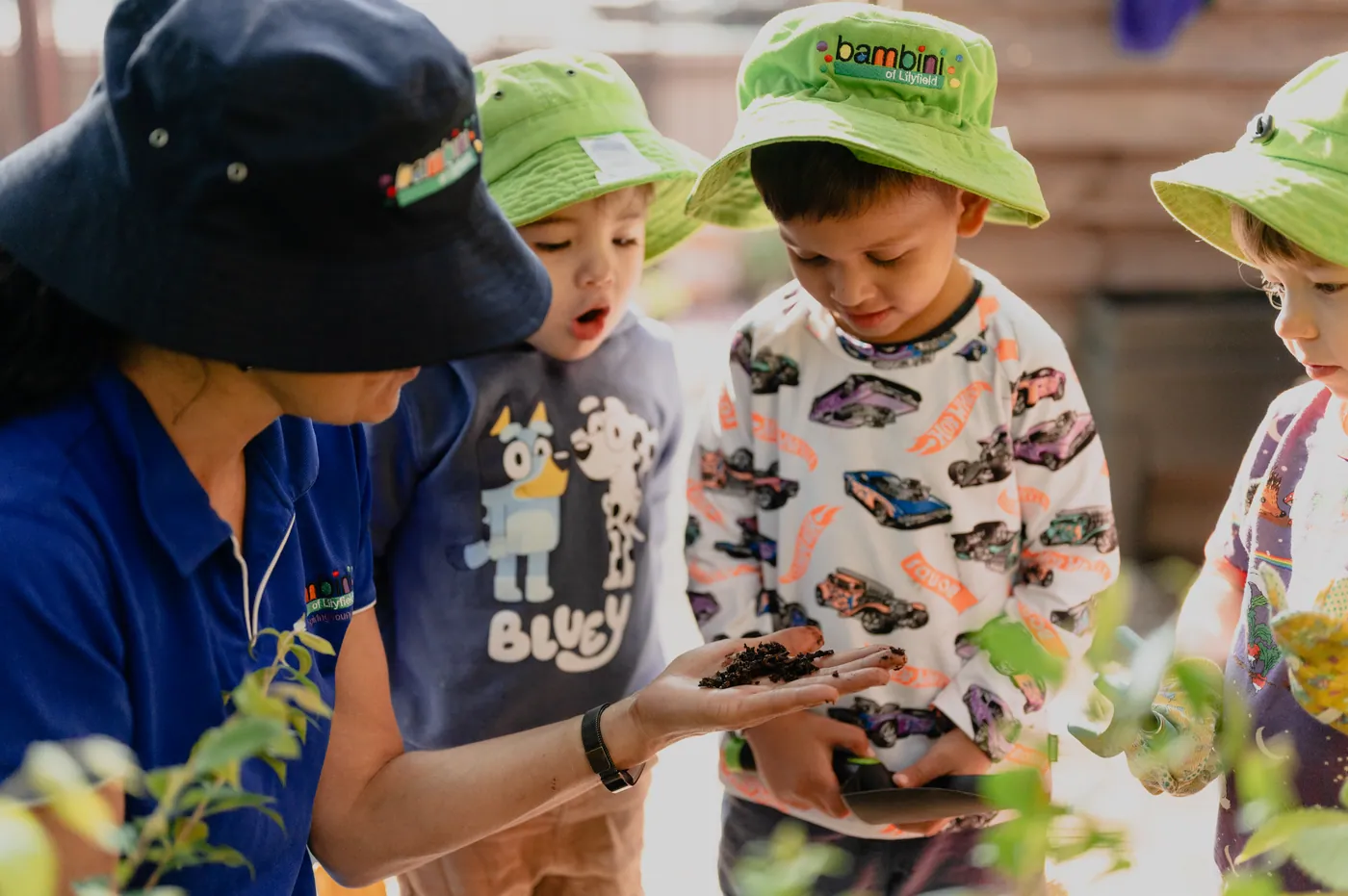 An educator holding a handful of dark soil in her palm while three children in Bambini hats lean in to look at it