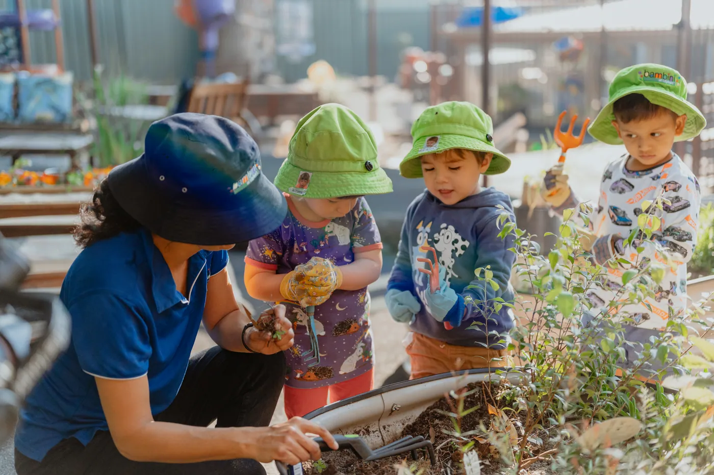 An educator kneeling at the raised garden bed with three children in Bambini hats, two of them wearing gardening gloves and holding trowels