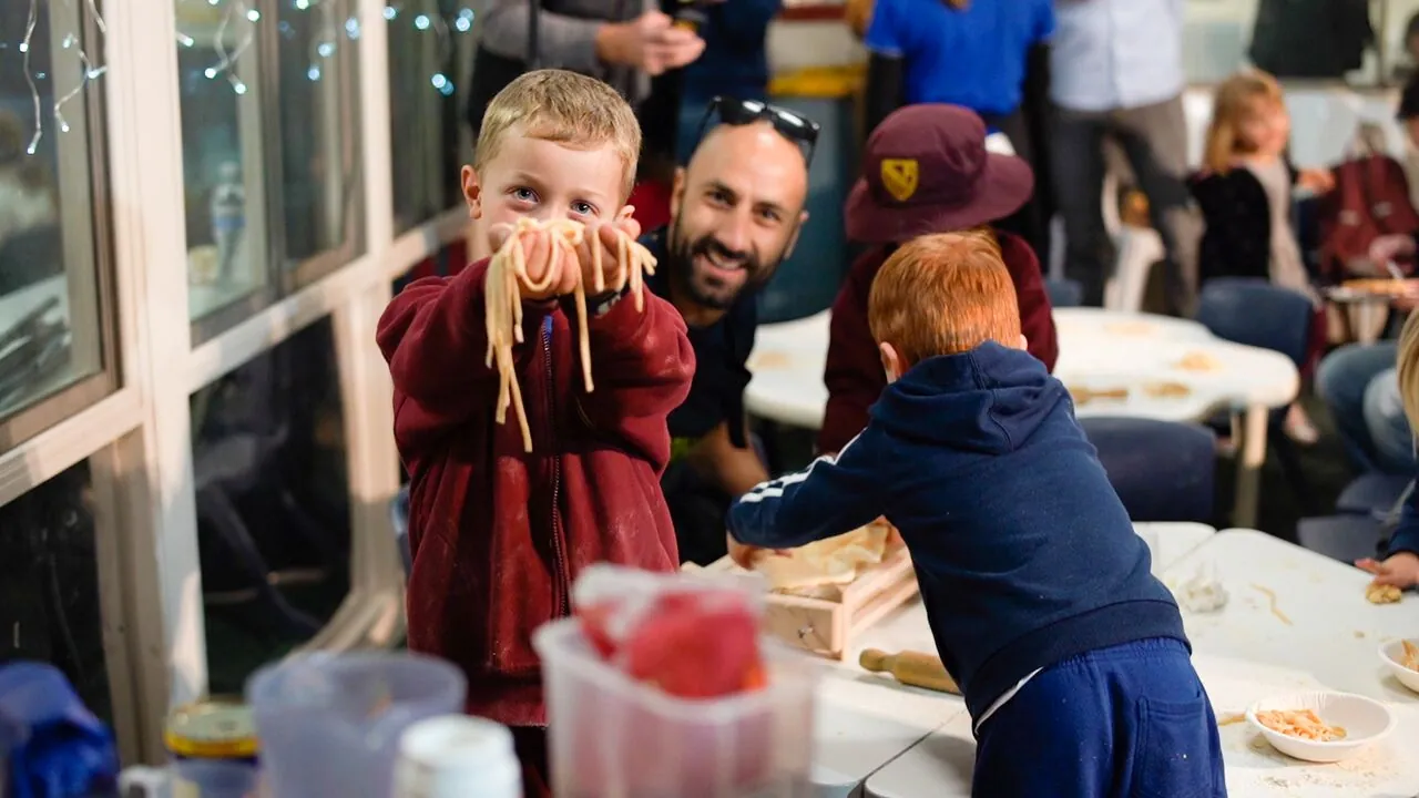 A families evening at the centre: a boy holding up strands of fresh pasta to the camera while his dad laughs behind him and another child rolls dough at the table