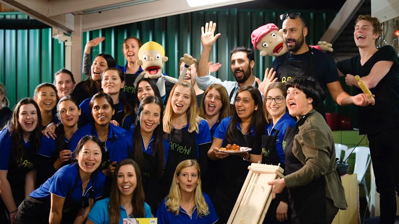 The Bambini team crowded together for a group photo at a community event, most in blue Bambini polos and black aprons, waving and holding up two puppets