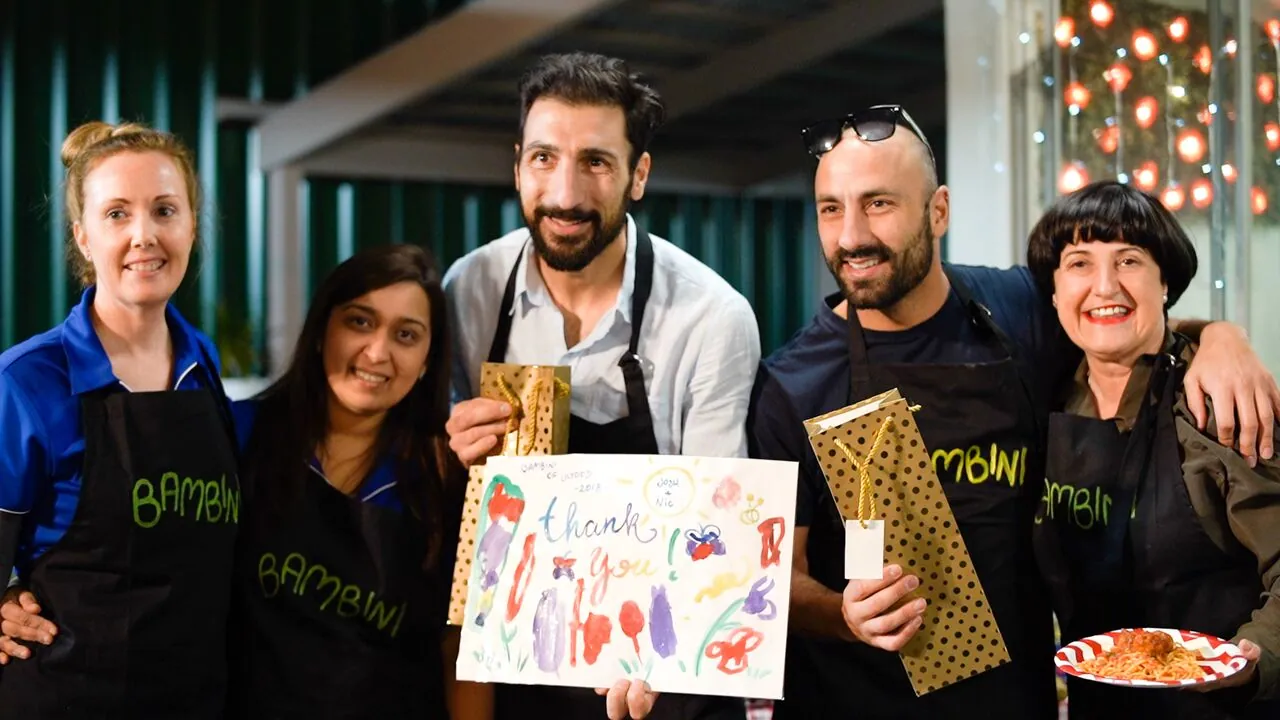 Five adults at an evening event, three of them in black Bambini aprons, holding up a child's painted thank you card and two gold gift bags, with a plate of spaghetti at the edge of the frame