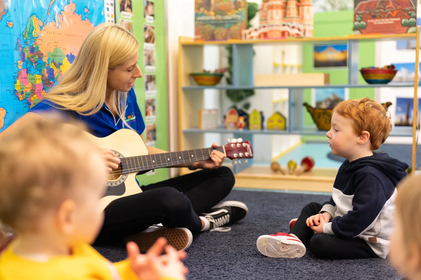 An educator sitting on the floor playing guitar while a toddler watches, with other children nearby