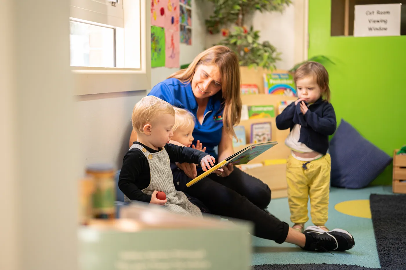 An educator sitting on the floor of the toddler room reading a picture book to two toddlers, while a third stands watching by the bookshelf