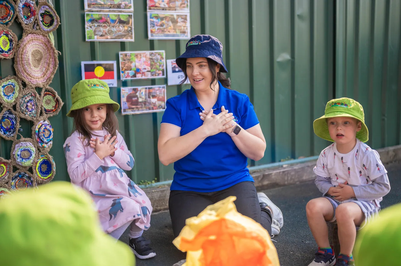 An educator in a blue Bambini polo and sun hat signing a song with two children in green Bambini hats, sitting outside against a green wall hung with photos and an Aboriginal flag