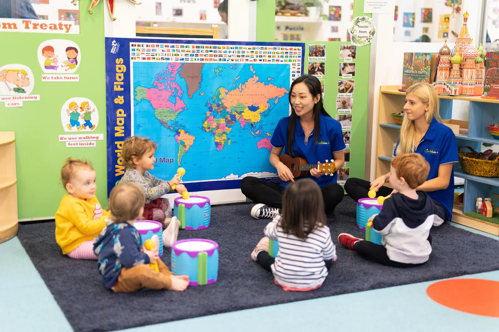Two educators, one playing a ukulele, sitting on the rug with toddlers and their drums in front of a large world map and flags poster