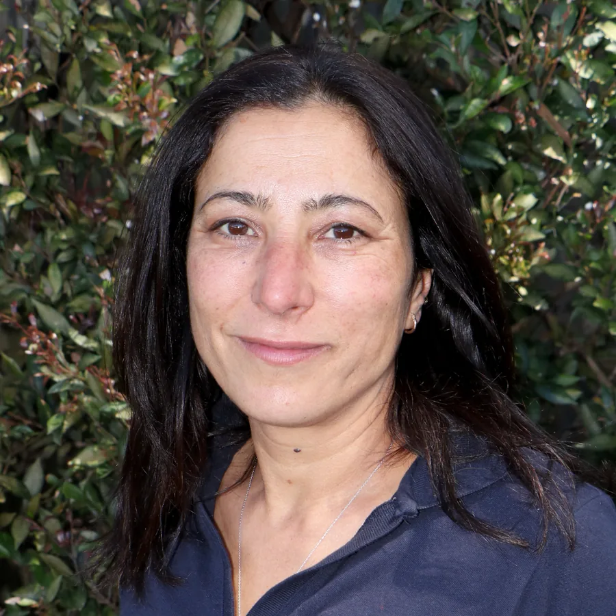 Anita smiling to camera, dark shoulder-length hair, in Bambini uniform in front of the centre’s green hedge