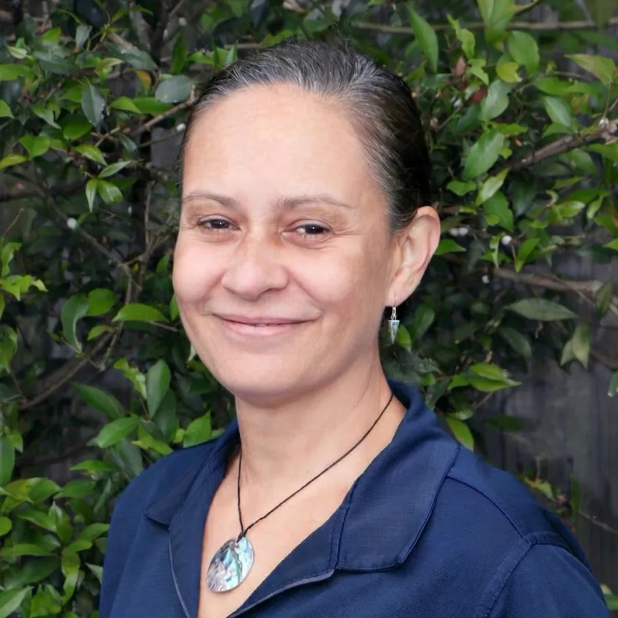 Carmel smiling to camera, dark hair tied back and a paua shell pendant, in Bambini uniform in front of the centre’s green hedge
