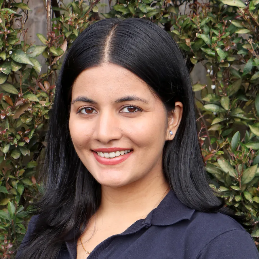 Deeya smiling to camera, long dark hair, in Bambini uniform in front of the centre’s green hedge