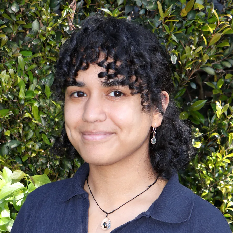 Georgia smiling to camera, dark curly hair, in Bambini uniform in front of the centre’s green hedge