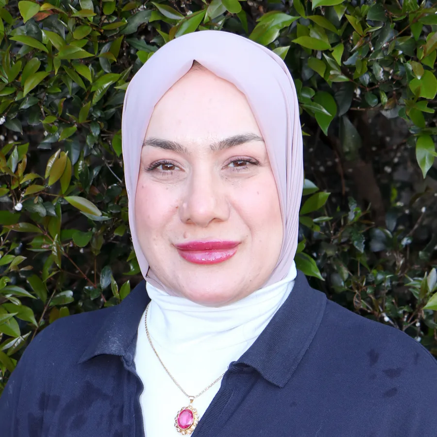 Hiba smiling to camera, wearing a pale lilac hijab, in Bambini uniform in front of the centre’s green hedge