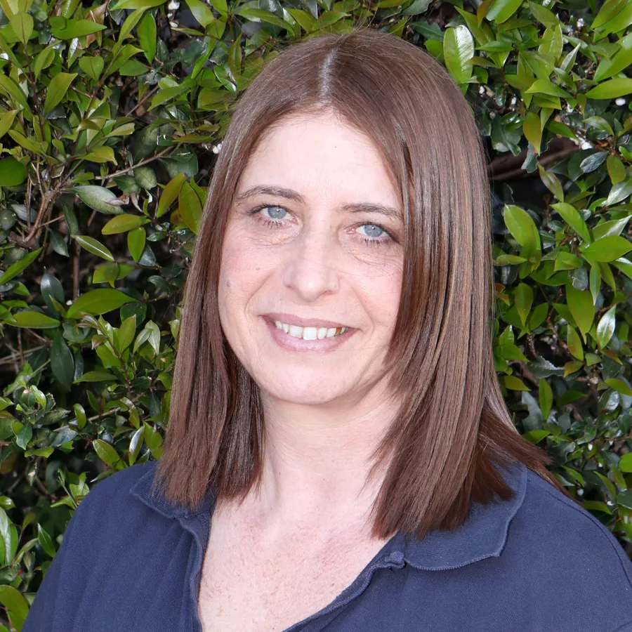 Jenny smiling to camera, shoulder-length brown hair, in Bambini uniform in front of the centre’s green hedge