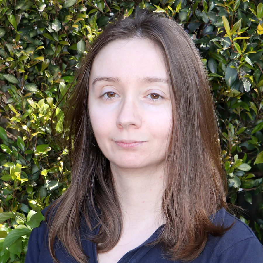 Patty looking to camera, long brown hair, in Bambini uniform in front of the centre’s green hedge