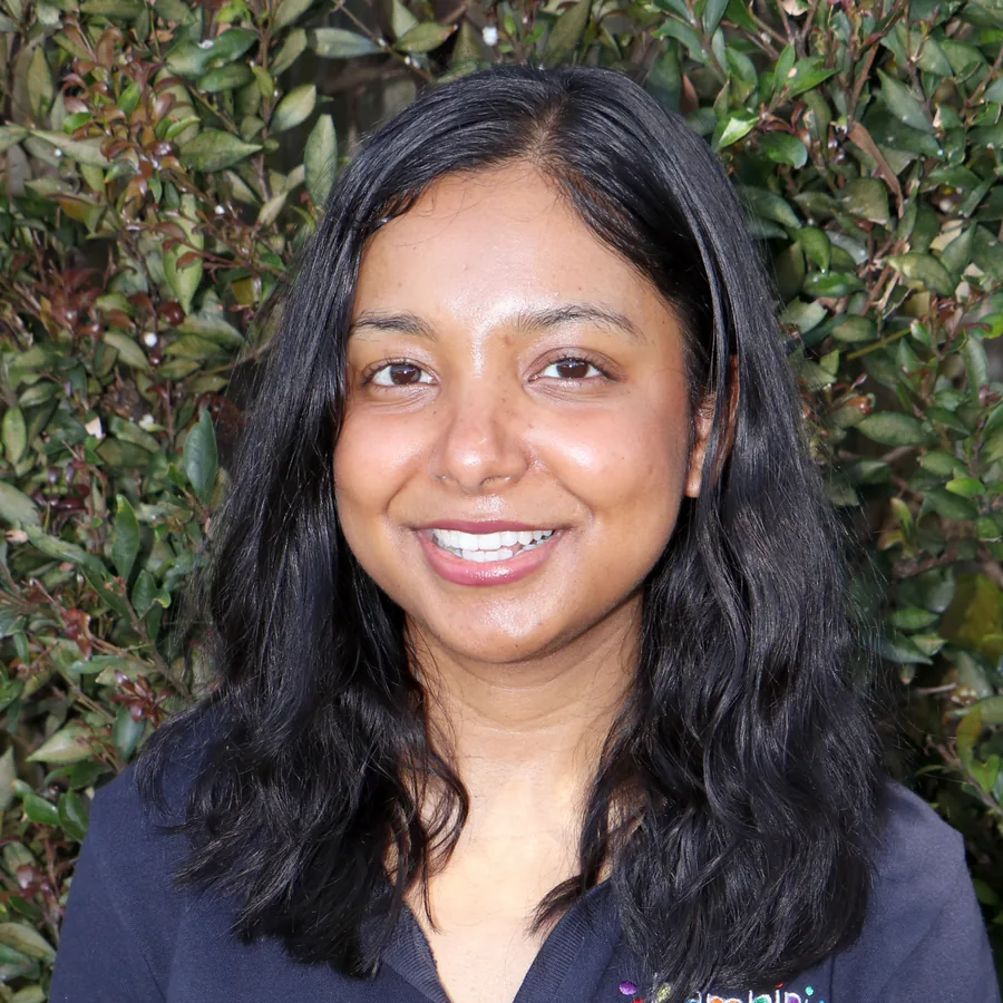 Pragati smiling to camera, long dark wavy hair, in Bambini uniform in front of the centre’s green hedge