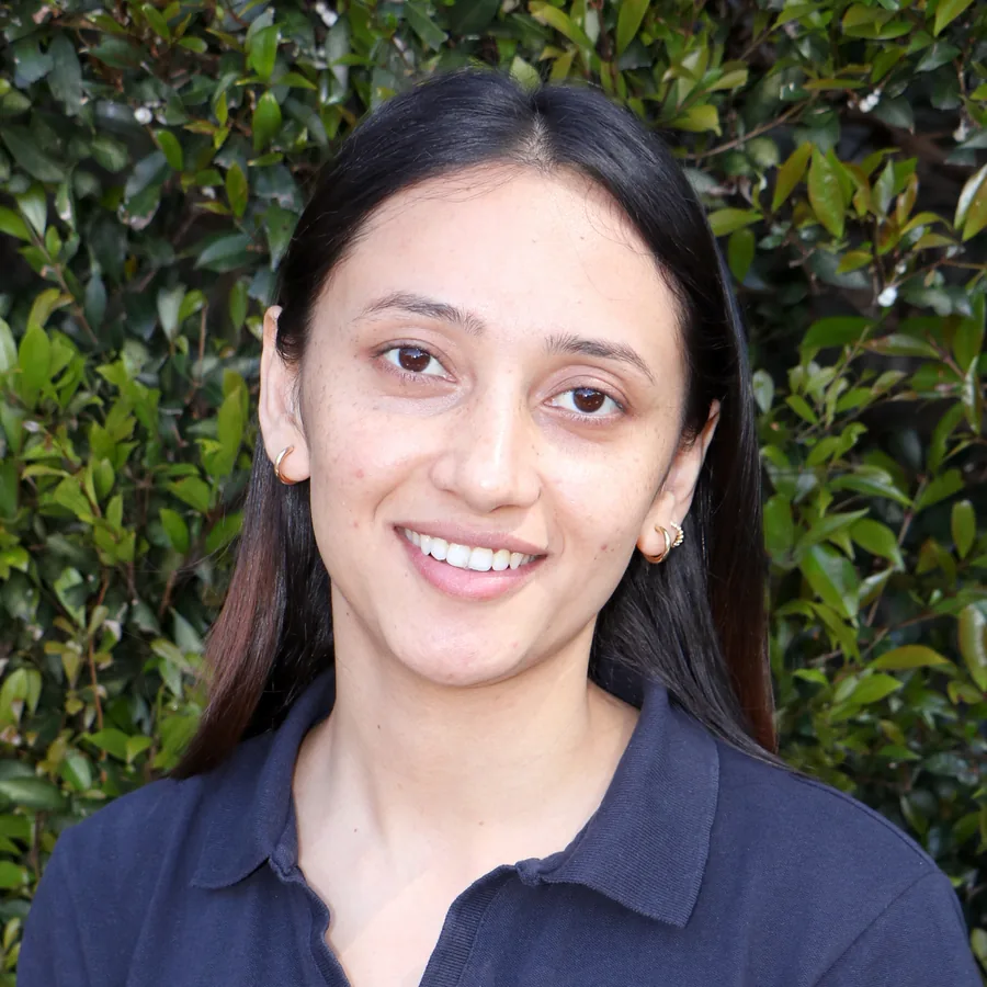 Susmita smiling to camera, long dark hair, in Bambini uniform in front of the centre’s green hedge